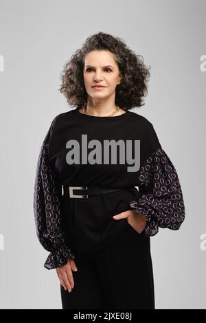 Femme âgée avec des cheveux bouclés luxuriants et un pantasuit noir posé en studio sur fond gris Banque D'Images