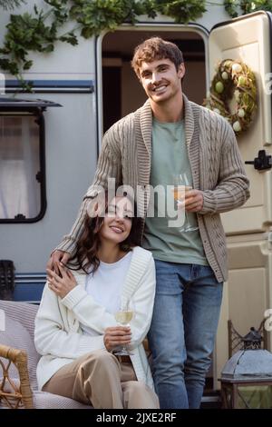 Jeune couple dans des cardigans chaleureux tenant le vin et souriant à l'appareil photo près de la camionnette de camping, image de stock Banque D'Images