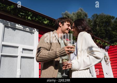 Vue à angle bas de jeune couple souriant dans cardigans tenant des verres de vin près de la maison à l'extérieur, image de stock Banque D'Images