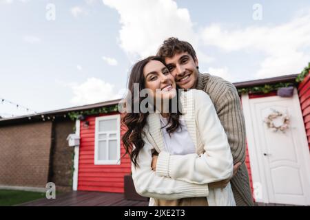 Couple gai dans cardigans embrassant et regardant la caméra près de la maison à l'extérieur, image de stock Banque D'Images