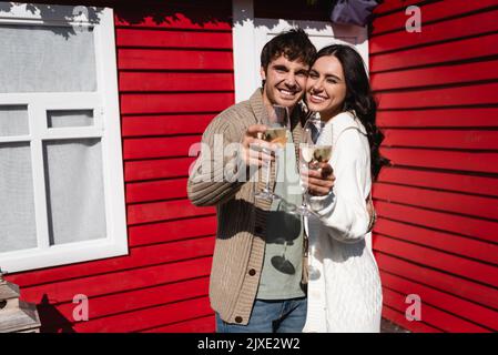 Couple positif en cardigans tenant des verres de vin près de la maison, image de stock Banque D'Images