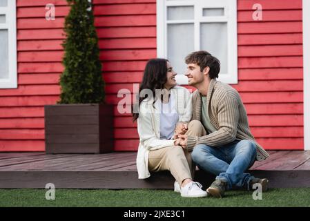 Vue latérale du jeune couple dans les cardigans chauds tenant les mains près de la maison floue, image de stock Banque D'Images