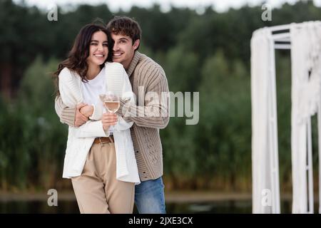 Couple souriant dans cardigans embrassant et tenant le vin à l'extérieur, image de stock Banque D'Images