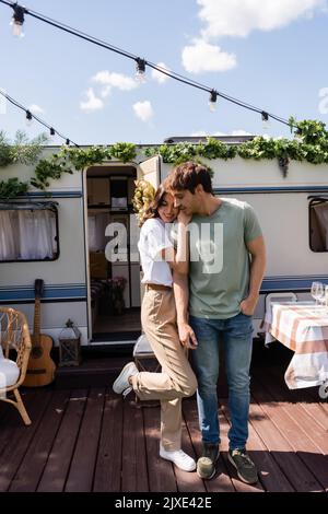 Femme souriante embrassant un petit ami sous des ampoules sur la terrasse près d'une camionnette de camping, image de stock Banque D'Images