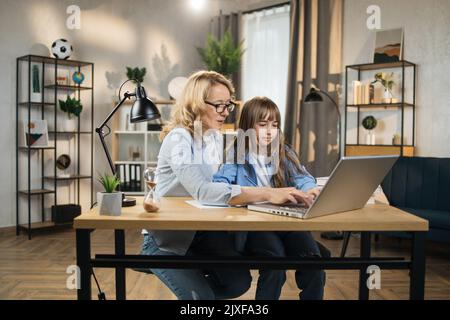 La jeune femme blonde de race blanche aide sa belle fille à faire ses devoirs en utilisant un ordinateur portable assis sur une table dans le salon à la maison. Banque D'Images