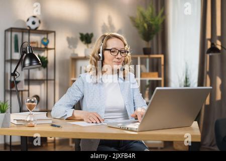 Portrait de belle confiante avec les cheveux blonds et sur les écouteurs femme intelligente portant des vêtements décontractés, assis à table devant l'ordinateur et de vérifier les informations dans la présentation sur Internet Banque D'Images