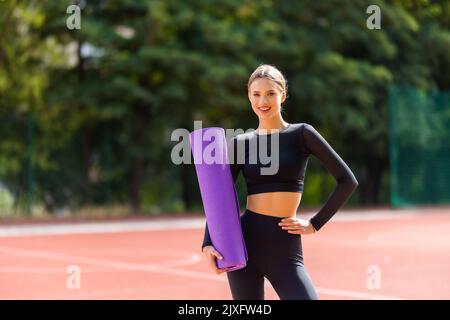 Joyeuse jeune fille caucasienne tenant le tapis de yoga se préparer pour l'entraînement debout à l'extérieur. Banque D'Images