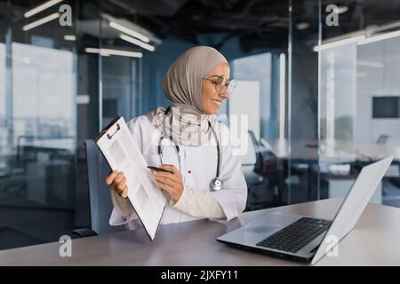 Médecin consultation en ligne avec le patient, femme musulmane dans le hijab travaillant à distance avec un ordinateur portable pour les appels vidéo, consultant les patients assis dans le bureau moderne de la clinique Banque D'Images