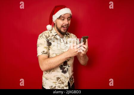 Jeune voyageur enthousiaste touriste homme dans les vêtements d'été, santa Hat utilisant le téléphone portable dactylographiant sms Banque D'Images