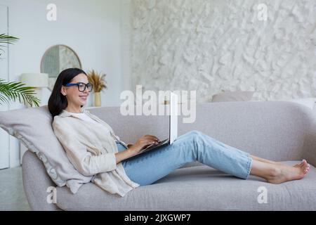 Travail en ligne. Jeune femme d'affaires en lunettes, freelance travaillant à la maison sur ordinateur portable. Allongé confortablement sur le canapé en jeans et en chemise, dactylographiant Banque D'Images