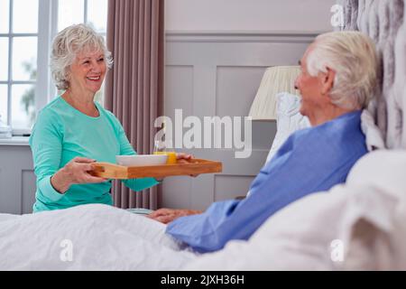 Couple Senior à la maison avec une femme apportant le petit déjeuner Senior Man au lit sur plateau Banque D'Images