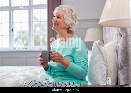 Femme sénior à la maison portant un pyjama assis sur le lit avec une tasse de thé du matin Banque D'Images