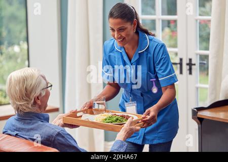 Femme de soins en uniforme apporter un repas sur plateau à l'homme senior assis dans le salon à la maison Banque D'Images