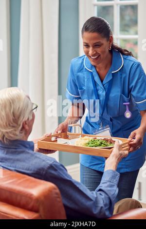 Femme de soins en uniforme apporter un repas sur plateau à l'homme senior assis dans le salon à la maison Banque D'Images