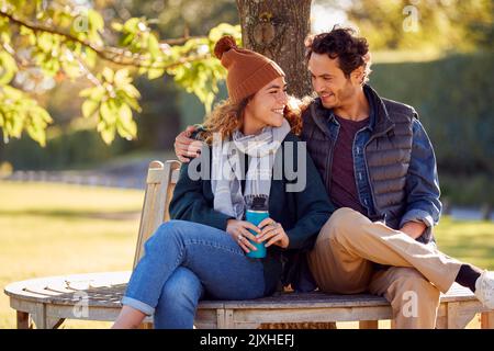 Couple souriant assis sur un banc dans le parc d'automne avec des tasses réutilisables Banque D'Images