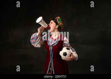 Une jeune femme surprise en tenue traditionnelle nationale ukrainienne crie au mégaphone isolé sur fond vintage sombre. Sport, Paris Banque D'Images