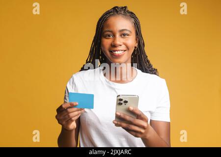 Image positive rêvant jolie femme posant isolée sur fond de mur jaune en utilisant le téléphone mobile tenant la carte de crédit Banque D'Images