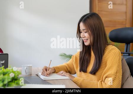 étudiante asiatique regardant la leçon en ligne et étudiant à distance. La jeune femme prend des notes, écoutant la conférence de l'enseignante sur appel vidéo.Video Banque D'Images