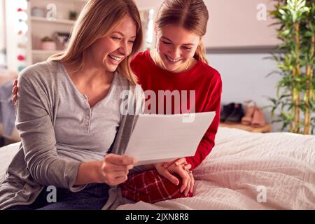 Fille adolescente souriante portant un pyjama regardant le rapport d'examen scolaire avec la mère dans la chambre Banque D'Images