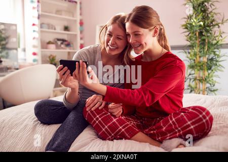 Mère avec fille adolescente portant un pyjama assis sur le lit posant pour Selfie sur le téléphone portable Banque D'Images