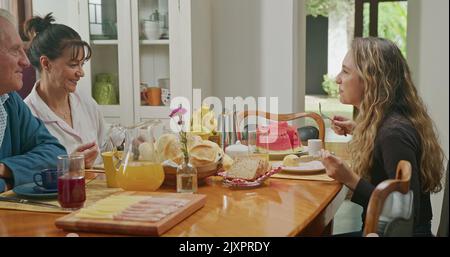 Un couple heureux prenant le petit déjeuner avec une fille adolescente assise à la table de la cuisine. Le père parle et la mère sert du jus à la fille Banque D'Images