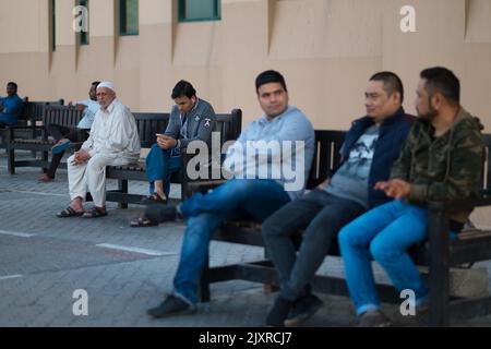 Des hommes de tous âges et de toutes races portant des vestes légères s'assoient sur une série de bancs par temps froid. Un vieil homme vêt de vêtements pakistanais traditionnels regarde la caméra. Banque D'Images
