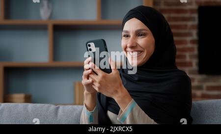 Souriant arabe musulman islamique 30s femme utilisatrice du foulard hijab noir utilisant le téléphone à la maison assis sur un canapé fille moyen-orientale tenant mobile Banque D'Images