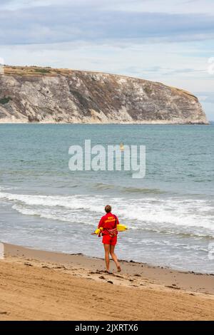 RNLI sauveteur marchant seul sur la plage de sable à swanage sur la côte du purbeck dorset avec des falaises en arrière-plan, institution nationale royale de sauvetage. Banque D'Images