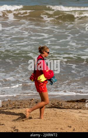 RNLI Lifeguard marchant le long de la plage de sable à wareham dans dorset, maître-nageur vêtu d'uniforme rouge marchant le long de la plage de sable transportant le flotteur de sauvetage. Banque D'Images