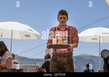 Un serveur portant du lederhosen sert un plateau de boissons aux convives dans un restaurant alpin des Dolomites italiens. Banque D'Images