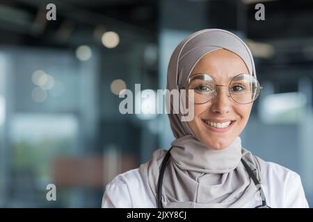 Portrait en gros plan d'une femme musulmane médecin en hijab gris, femme médecin souriant et regardant l'appareil photo, femme travailleuse en lunettes et blanc manteau médical et des lunettes travaillant dans le bureau moderne de la clinique Banque D'Images