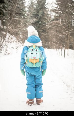 Garçon de six ans debout avec son dos à la caméra. Enfant avec sac à dos amusant dans la forêt d'hiver pleine de neige. Enfant vêtu de couleurs vives. Banque D'Images