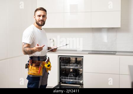 Jeune militaire moderne en costume de travailleur lors de la réparation du lave-vaisselle sur la cuisine domestique Banque D'Images