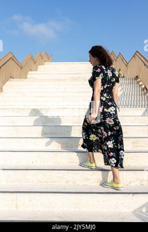 Une jeune fille de vingt ans vêtue d'une robe noire ornée de fleurs jaunes monte les escaliers en marbre Banque D'Images