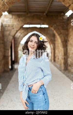 Une fille de brunette pensive aux cheveux longs et bouclés dans une chemise en lin blanc avec une bande bleue, un Jean bleu et des baskets vertes avec une fleur de plumeria blanche Banque D'Images