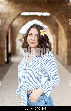 Une fille de brunette pensive aux cheveux longs et bouclés dans une chemise en lin blanc avec une bande bleue, un Jean bleu et des baskets vertes avec une fleur de plumeria blanche Banque D'Images