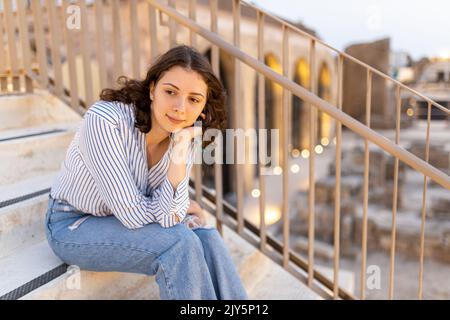 Une fille de brunette pensive aux cheveux longs et bouclés dans une chemise en lin blanc avec une bande bleue, un Jean bleu et des baskets vertes avec une fleur de plumeria blanche Banque D'Images