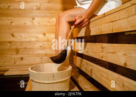 main de femme caucasienne inconnue dans le sauna spa prenant l'eau du seau avec une cuillère en bois pour verser sur les pierres chaudes Banque D'Images