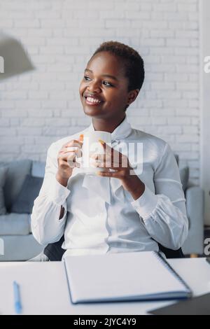 Une femme d'affaires africaine rêveuse qui apprécie une tasse de café, se reposer après le travail. Penser à un projet réussi, à un profit futur, sourire, rire, se sentir Banque D'Images