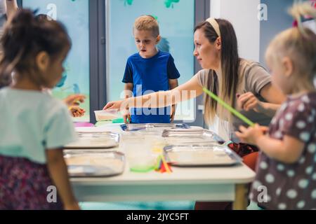 Jeu sensoriel à l'école maternelle multi-culturelle. Les tout-petits avec leur professeur jouant avec des pailles rayées et la peinture au lait, en utilisant la coloration de nourriture, le lait, le papier aquarelle, et les plateaux. Activité créative pour les enfants pour le développement du cerveau. Peinture au doigt. Photo de haute qualité Banque D'Images