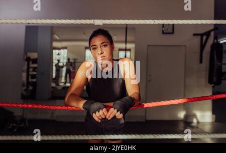 Boxeur féminin confiant regardant la caméra tout en s'appuyant sur les cordes dans un anneau de boxe. Une jeune femme sportive s'entraîne à la salle de gym. Banque D'Images