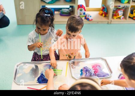 Jeu sensoriel à l'école maternelle multi-culturelle. Les tout-petits avec leur professeur jouant avec des pailles rayées et la peinture au lait, en utilisant la coloration de nourriture, le lait, le papier aquarelle, et les plateaux. Activité créative pour les enfants pour le développement du cerveau. Peinture au doigt. Photo de haute qualité Banque D'Images