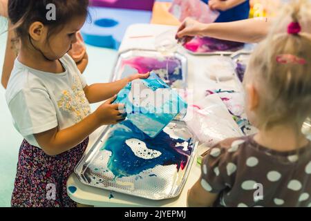 Jeu sensoriel à l'école maternelle multi-culturelle. Les tout-petits avec leur professeur jouant avec des pailles rayées et la peinture au lait, en utilisant la coloration de nourriture, le lait, le papier aquarelle, et les plateaux. Activité créative pour les enfants pour le développement du cerveau. Peinture au doigt. Photo de haute qualité Banque D'Images