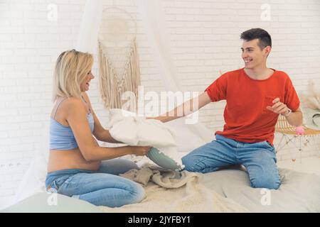 Charmant couple européen heureux - femme blonde enceinte et son mari aux cheveux sombres - assis sur un lit ou un canapé, riant et se battant avec des oreillers. Concept de mariage heureux. Photo de haute qualité Banque D'Images