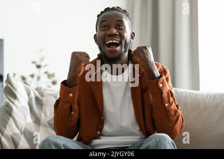 Joyeux homme noir en train de gester OUI dans la joie et crier, en se sentant heureux, en applaudant pour l'équipe de sport, assis sur le canapé à la maison Banque D'Images