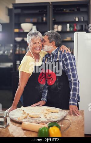 Un couple heureux et mûr qui partage un moment de tendresse avec un baiser tout en cuisinant ensemble à la maison. Banque D'Images