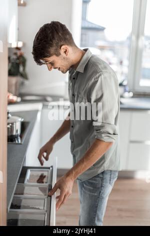 Jeune homme ouvre le réfrigérateur encastré dans sa cuisine. Banque D'Images