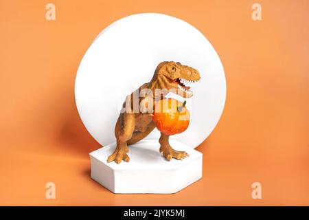 Jouet dinosaure Tyrannosaurus tenant le potiron dans ses pattes, sur le podium blanc isolé sur fond orange carte de voeux de vacances Joyeux jour d'Halloween, Bonjour Banque D'Images