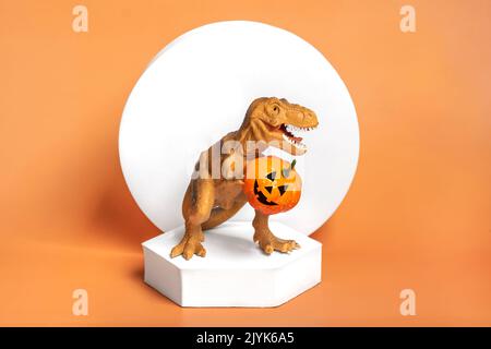 Jouet dinosaure Tyrannosaurus tenant le potiron dans ses pattes, sur le podium blanc isolé sur fond orange carte de voeux de vacances Joyeux jour d'Halloween, Bonjour Banque D'Images