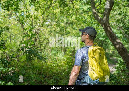 Vue latérale d'un randonneur mâle portant une casquette de lunettes de soleil et un sac à dos qui se promette dans une forêt britannique lors d'une journée ensoleillée d'été. Banque D'Images